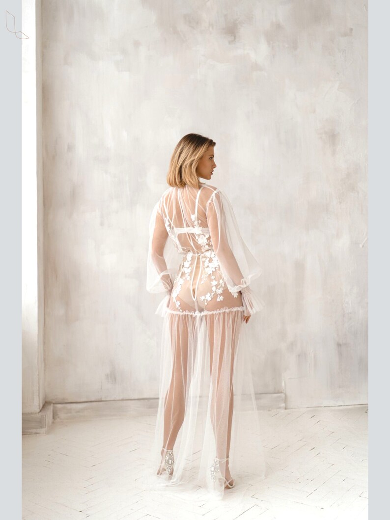 Pu&ograve; includere: Un abito maxi bianco in tulle trasparente con ricami floreali e una gonna lunga e fluida. L'abito ha un corpetto aderente e maniche lunghe con un dettaglio arricciato sulle spalle.
