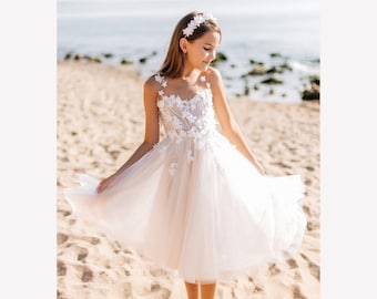 Puede incluir: Una niña pequeña con un vestido de tul blanco con bordados florales y una diadema de flores blancas. Está de pie en una playa de arena.