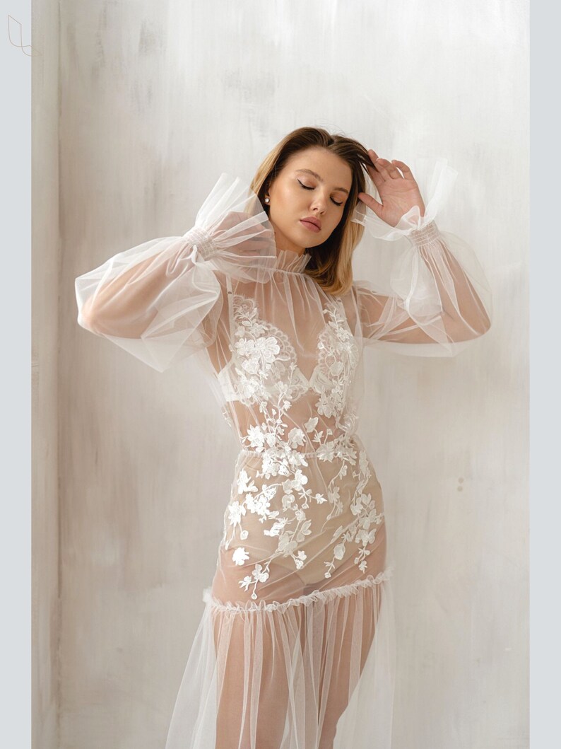 Pu&ograve; includere: Un abito bianco trasparente con maniche lunghe e un design in pizzo floreale. L'abito ha un collo alto e un orlo arricciato.