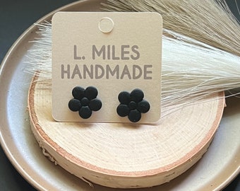 Puede incluir: Un par de pendientes negros en forma de flor sobre un expositor de madera con una tarjeta que dice "L. MILES HANDMADE".
