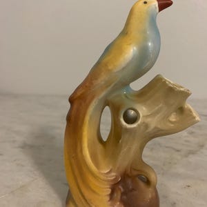 May include: A ceramic bird figurine with a yellow, blue, and brown glaze. The bird is perched on a tree branch with a hole in the center. The figurine is a vintage style and would be a great addition to any home decor.