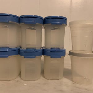 Puede incluir: Un conjunto de recipientes de plástico transparente para guardar alimentos con tapas azules. Los recipientes son rectangulares y están apilados. También hay dos recipientes redondos más pequeños con tapas grises.