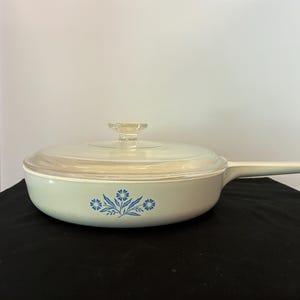 May include: A white ceramic skillet with a long handle and a clear glass lid. The exterior of the pan features a blue floral pattern consisting of three stylized flowers with leaves centered on the side of the round cooking vessel.