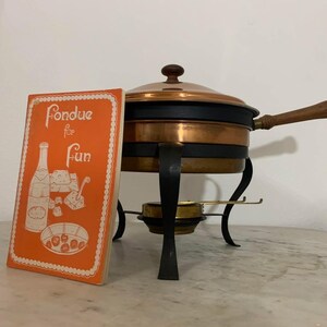 May include: A copper and black fondue pot with a wooden handle and a matching lid. A vintage cookbook titled "Fondue for Fun" is propped up next to the pot. The cookbook is orange with white illustrations.
