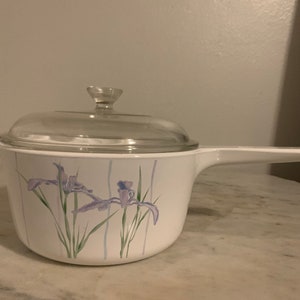 May include: White ceramic saucepan with a glass lid and a floral design featuring purple irises. The handle is white and the saucepan has a white background with green and blue accents.