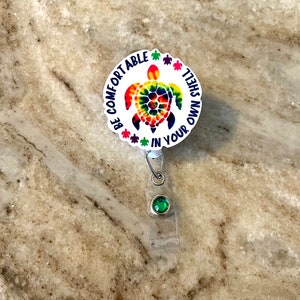 May include: A white round badge holder with a colorful tie-dye turtle design. The badge holder has the text "Be comfortable in your own shell" around the turtle. The badge holder has a green gem on the bottom.