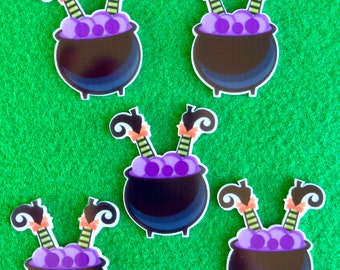 May include: Five Halloween-themed stickers featuring a black cauldron with purple bubbling liquid and witch legs. The witch legs are black with orange bows and green and yellow striped stockings. The stickers are on a green background.