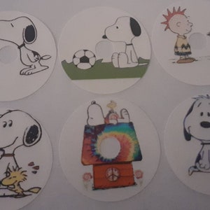Peut inclure: Six autocollants circulaires blancs avec Snoopy et d'autres personnages de Peanuts. Les illustrations incluent Snoopy avec une fleur, jouant au football, et Snoopy avec Woodstock.