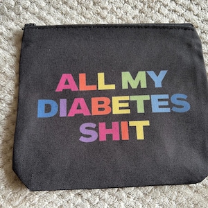 Könnte beinhalten: Schwarze Reißverschlusstasche mit dem Text "ALL MY DIABETES SHIT" in Regenbogenfarben. Die Tasche ist rechteckig und scheint aus einem strapazierfähigen Stoff zu sein. Der Hintergrund ist eine strukturierte, hellfarbige Oberfläche.