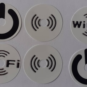 Puede incluir: Seis pegatinas redondas blancas con símbolos negros. Tres pegatinas tienen un símbolo de botón de encendido negro. Tres pegatinas tienen un símbolo Wi-Fi negro con el texto "Wi Fi" en negro.