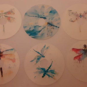 May include: Six white circular coasters, each with a watercolour dragonfly design. The dragonflies are in various colours, including blue, pink, and orange. The coasters have a hole in the centre.