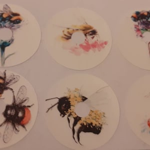 May include: Six round stickers featuring different bee designs. Each sticker depicts a bee with watercolour-style illustrations of flowers and leaves. The bees are yellow and black with brown and white accents.