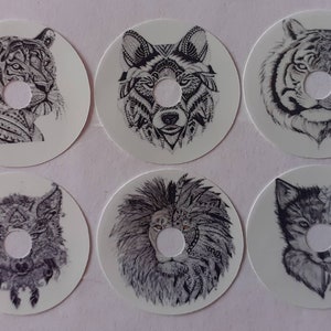 Può includere: Sei adesivi rotondi bianchi con disegni dettagliati in bianco e nero di volti di animali. Gli animali sono una tigre, un lupo, un leone e un lupo con un motivo geometrico. Ogni adesivo ha un foro rotondo al centro.
