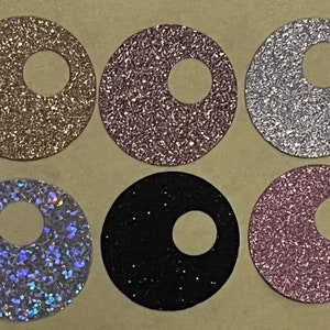 May include: Six round, glittery, adhesive stickers with a hole in the center. The stickers are in various colors, including gold, silver, black, pink, and iridescent.