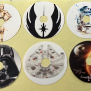 Puede incluir: Seis pegatinas redondas blancas con diferentes personajes e imágenes de Star Wars. Las pegatinas tienen un agujero en el centro. Los personajes incluyen Darth Vader, R2-D2, C-3PO, el Halcón Milenario, el símbolo de la Alianza Rebelde y una escena de la Estrella de la Muerte.
