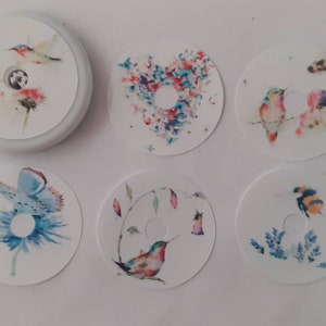 May include: Six round, white, paper stickers with watercolour designs featuring hummingbirds, bees, and flowers. The stickers have a hole in the centre.
