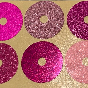 Puede incluir: Seis pegatinas circulares con un agujero en el centro, en tonos rosa y magenta. Las pegatinas tienen acabados brillantes y texturizados. Las pegatinas están dispuestas sobre un fondo beige claro.
