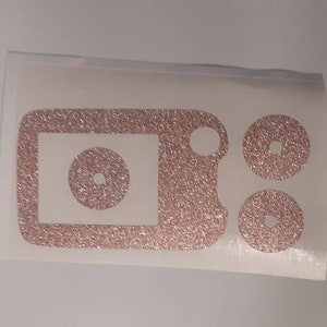 May include: A set of three rose gold glitter vinyl decals for a phone or other device. The decals are in the shape of a rectangle with a circle inside, and two smaller circles.