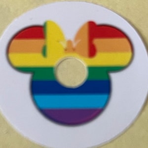 Puede incluir: Tres pegatinas redondas blancas con un diseño de cabeza de Mickey Mouse arcoíris. La cabeza de Mickey Mouse está delineada en negro y tiene un patrón de rayas arcoíris.