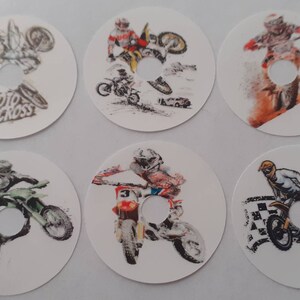 May include: Six round stickers featuring motocross riders in various poses. The stickers are white with a hole in the centre. The riders are wearing helmets and riding dirt bikes. The stickers have a motocross theme.