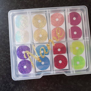 May include: A clear plastic storage box with 12 compartments, each containing a round, glittery sticker. The stickers are in various colours, including purple, blue, red, green, and yellow. The word "Name" is written in gold glitter across the top of the box.