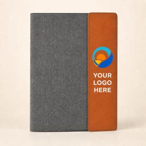 May include: A rectangular notebook featuring a dark gray fabric cover on the left side and a smooth tan leather-like vertical strip on the right. The tan section displays a circular orange and blue logo above the white text YOUR LOGO HERE.