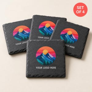 May include: A set of four square black slate coasters with rough, textured edges. Each coaster features a circular graphic of a colourful mountain landscape at sunset with a yellow sun, blue peaks, and red sky, with the text YOUR LOGO HERE printed below.