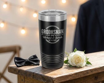 May include: Black insulated tumbler with a silver rim, engraved with the word "GROOMSMAN", names, and a date. A black bow tie and a white rose are on a wooden surface. Decorative lights are in the background.