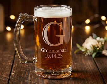 May include: A clear glass beer mug filled with beer and foam. The mug has a handle and is engraved with a large letter "G", the word "Groomsman", and the date "10.14.23". The mug sits on a wooden surface.