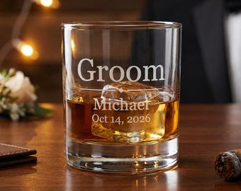 May include: A clear glass tumbler with the word "Groom", a name, and a date etched into the glass. The glass is filled with amber liquid and a large ice cube. A lit cigar and a small bouquet are in the background.