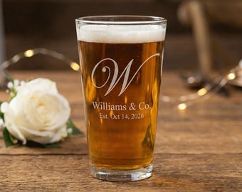 May include: A clear pint glass filled with beer, featuring a white foam head. The glass is etched with a large script "W" and the text "Williams & Co. Est. Oct 14, 2026". A white rose and greenery are in the background.