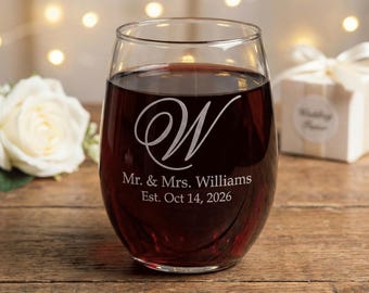 May include: A stemless wine glass filled with red wine, engraved with a stylized "W" and the text "Mr. & Mrs. Williams Est. Oct 14, 2026". A white rose and a gift box with a bow are in the background. The glass sits on a wooden surface.