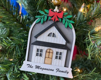 May include: A wooden Christmas ornament shaped like a house, hanging on a Christmas tree. The house has a gray roof and white siding, with a red bow and green leaves at the top. The text "The Magnuson Family" is written on the bottom.