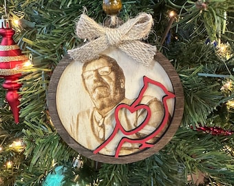 May include: A wooden Christmas ornament featuring a portrait of a person with glasses, laser-etched into the wood. A red cardinal design is layered over the portrait. The ornament is round, with a twine hanger and a burlap bow.
