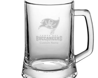 May include: A clear glass beer stein with a Tampa Bay Buccaneers logo and the text "Tampa Bay Buccaneers" and "Custom Name" etched on the front.