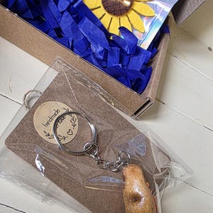 May include: A gift box with blue paper filler contains a felt sunflower and a keychain. The keychain has a brown potato-shaped charm and a tag that says "handmade with love".