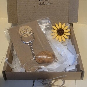 May include: A gift box containing a keychain with a brown stone, a yellow and brown sunflower pin, and a heart-shaped ornament. The box has the text "Focus on the journey not the destination".