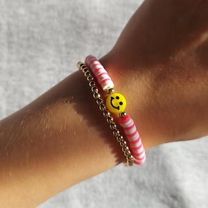 Puede incluir: Dos pulseras en una muñeca. Una pulsera está hecha de cuentas rayadas de color rosa y blanco. La otra pulsera está hecha de cuentas doradas y tiene un dije de carita sonriente amarilla.