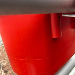 Może przedstawiać: Jaskrawoczerwony plastikowy pojemnik o gładkim, zaokrąglonym korpusie z poziomym szarym paskiem u g&oacute;ry. Pojemnik posiada cylindryczny punkt mocowania z boku oraz metalowy mechanizm zawiasowy widoczny na prawej krawędzi.
