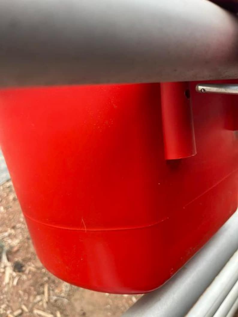Może przedstawiać: Jaskrawoczerwony plastikowy pojemnik o gładkim, zaokrąglonym korpusie z poziomym szarym paskiem u g&oacute;ry. Pojemnik posiada cylindryczny punkt mocowania z boku oraz metalowy mechanizm zawiasowy widoczny na prawej krawędzi.