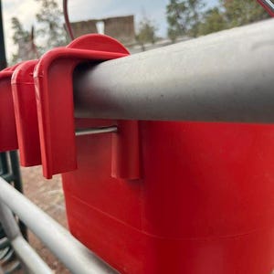 Może przedstawiać: Jaskrawoczerwony plastikowy pojemnik z solidnym mechanizmem haczykowym, zaprojektowany do bezpiecznego zawieszenia na poziomej metalowej poprzeczce. Pojemnik ma gładką, jednolitą powierzchnię oraz cienki metalowy uchwyt przymocowany do g&oacute;rnej krawędzi.