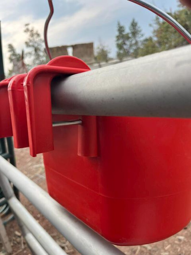 Może przedstawiać: Jaskrawoczerwony plastikowy pojemnik z solidnym mechanizmem haczykowym, zaprojektowany do bezpiecznego zawieszenia na poziomej metalowej poprzeczce. Pojemnik ma gładką, jednolitą powierzchnię oraz cienki metalowy uchwyt przymocowany do g&oacute;rnej krawędzi.