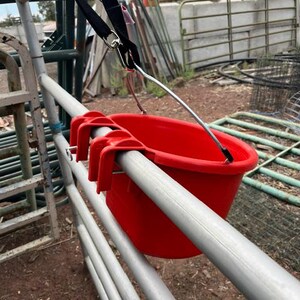 Może przedstawiać: Jaskrawoczerwone plastikowe wiadro z trzema zintegrowanymi haczykami na krawędzi, zawieszone na metalowym pręcie za pomocą czarnego regulowanego paska ze srebrnym metalowym klipsem oraz cienkiego metalowego wspornika przymocowanego do krawędzi wiadra.