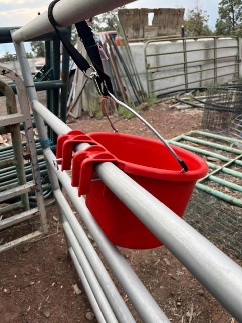 Może przedstawiać: Jaskrawoczerwone plastikowe wiadro z trzema zintegrowanymi haczykami na krawędzi, zawieszone na metalowym pręcie za pomocą czarnego regulowanego paska ze srebrnym metalowym klipsem oraz cienkiego metalowego wspornika przymocowanego do krawędzi wiadra.