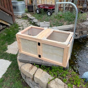 May include: A wooden fish trap with two compartments, each covered with wire mesh. The trap is made of light-colored wood and sits on a stone wall near a pond. A metal support arm extends above the trap.