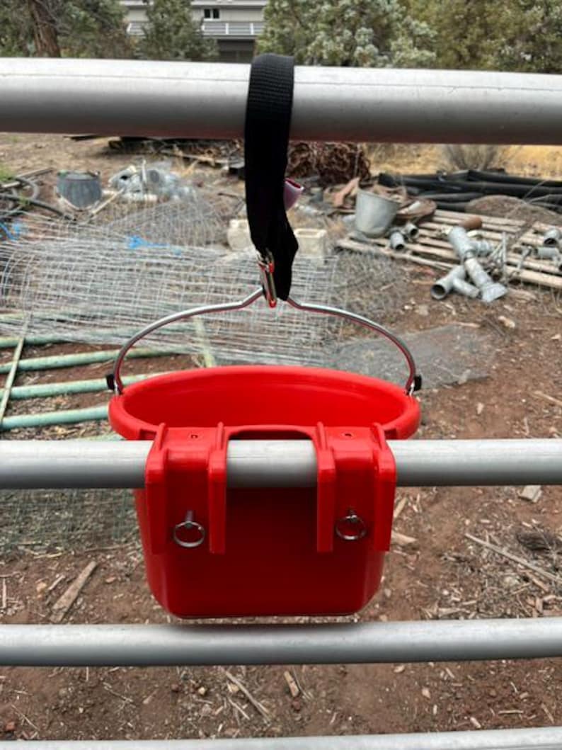 Może przedstawiać: Jaskrawoczerwone plastikowe wiadro z metalowym uchwytem i dwoma zintegrowanymi haczykami do montażu. Wiadro jest zawieszone na poziomej metalowej poprzeczce za pomocą czarnego nylonowego paska z metalowym klipsem i posiada dwa małe srebrne pierścienie.