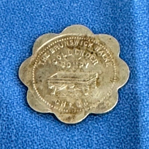 Könnte beinhalten: Ein silberfarbener Token mit dem Text "The Brunswick Balke Collender Company" und einer Darstellung eines Sarges.