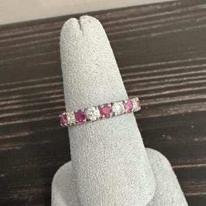 May include: A silver-toned eternity band ring featuring an alternating pattern of round-cut, deep pink rubies and clear, sparkling white diamonds set in a continuous row. The metal band has a polished finish, showcasing the gemstones clearly.