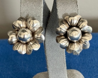 May include: A pair of silver flower-shaped stud earrings. The earrings are on a gray display stand.