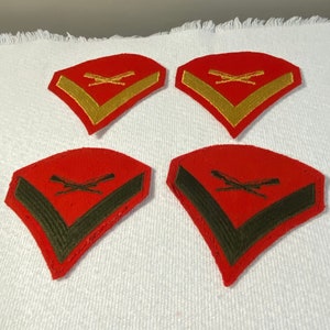 May include: Four red military rank insignia patches with gold and green embroidered details. The patches have a chevron shape with crossed rifles in the center.